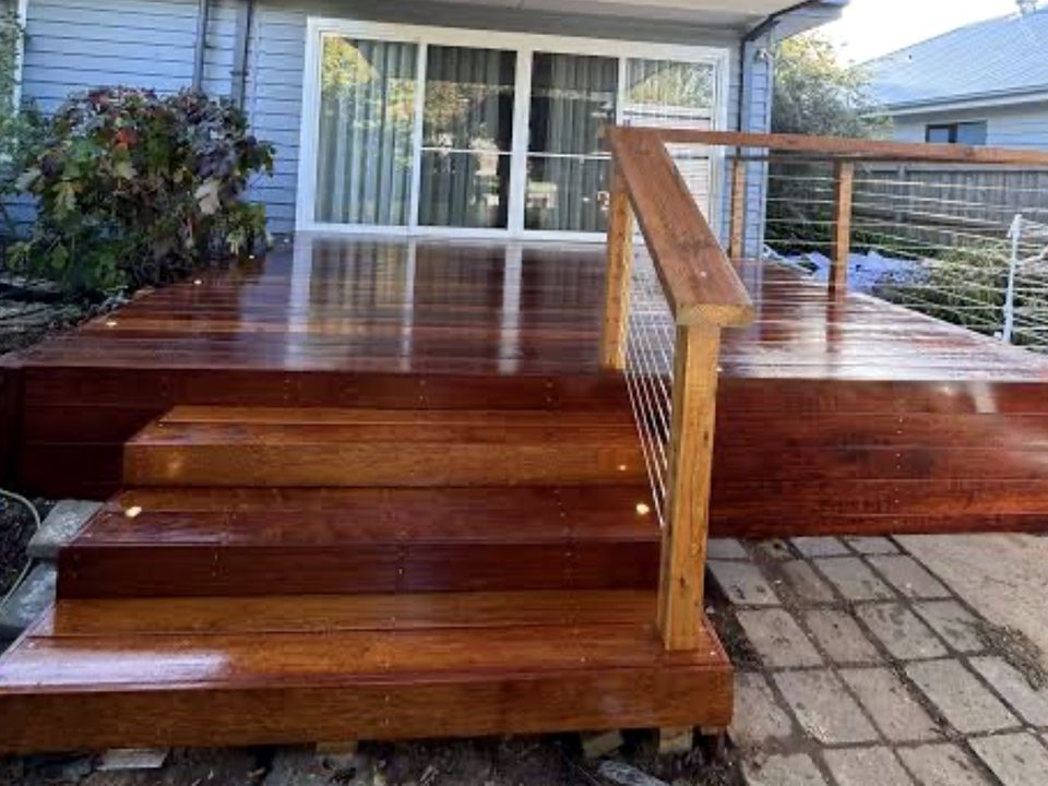 A wet, freshly-oiled red-stained hardwood deck with wide entry steps and a timber-and-wire handrail against a blue weatherboard home.