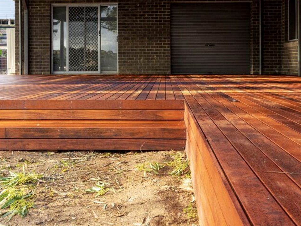 A low-profile hardwood deck with a wide step-down edge in front of a brick home, warm sunlight raking across the boards.