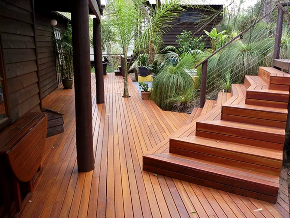 A wide oiled hardwood deck wrapping a timber-clad home, with cascading corner stairs, a wire balustrade and tropical planting.