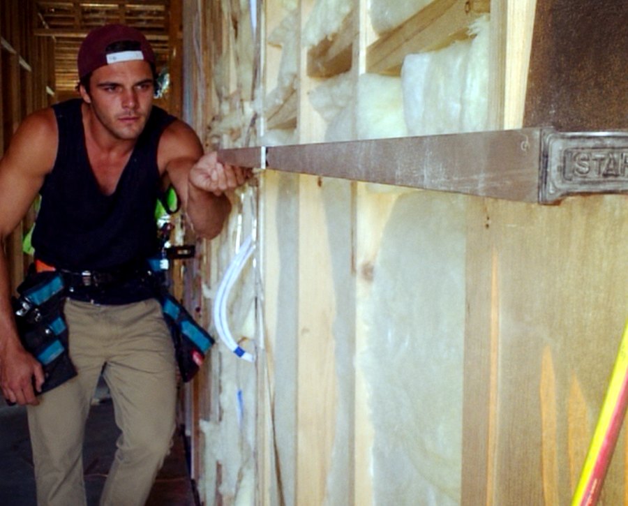 A younger Corey with his tool belt, sighting down a straightedge to check wall framing is true, insulation visible behind.